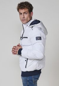 Chaqueta de plumas blanca con capucha, que presenta cremalleras negras y detalles en azul marino. Puños y dobladillo acanalados, parche del logo en la manga izquierda.