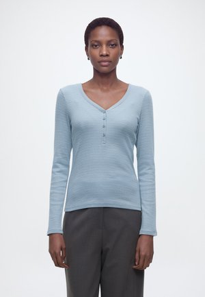 Femme aux cheveux courts portant un haut henley côtelé à manches longues bleu clair et un pantalon gris foncé, debout devant un fond blanc uni.