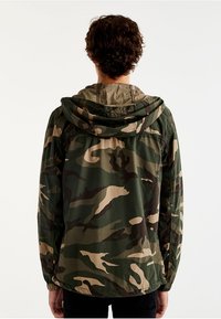 Giacca a vento camo in tonalità verde e tan con cappuccio regolabile, realizzata in un tessuto leggero e con vestibilità rilassata.
