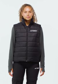 Young person with curly hair wearing black Adidas Terrex padded vest and matching black Terrex pants, standing against plain background.