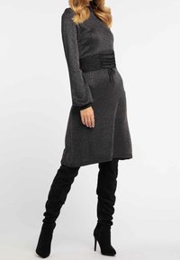 Femme dans une robe gris foncé à manches longues et paillettes, avec une taille froncée, associée à des bottes à talons aiguilles en suède noir montant jusqu'aux genoux.