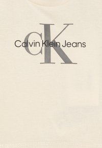 Tričko Calvin Klein Jeans v barvě off-white z bavlny s výrazným černým a šedým logem. Má kulatý výstřih a hladkou texturu.