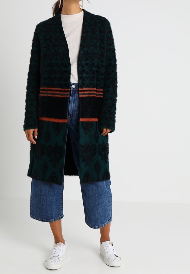 Lange, donkergroene cardigan met zwarte bloemmotieven en oranje strepen aan de zoom, gemaakt van zacht, gestructureerd materiaal, gecombineerd met jeans en sneakers.