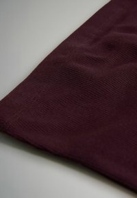 Detailní pohled na bordó žebrovanou látku s viditelným švem, rozloženou na světle šedém povrchu.