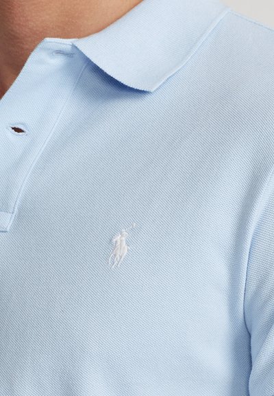 Lyseblå polo-shirt med tekstureret stof og et lille hvidt broderet logo af en hest og rytter på brystet.