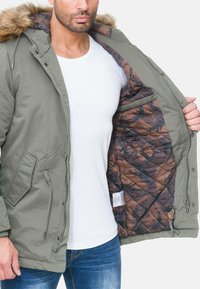 Parka verde oliva con cappuccio foderato in pelliccia, con interno trapuntato marrone e mimetico, tasche laterali e dettagli con cordino in vita.