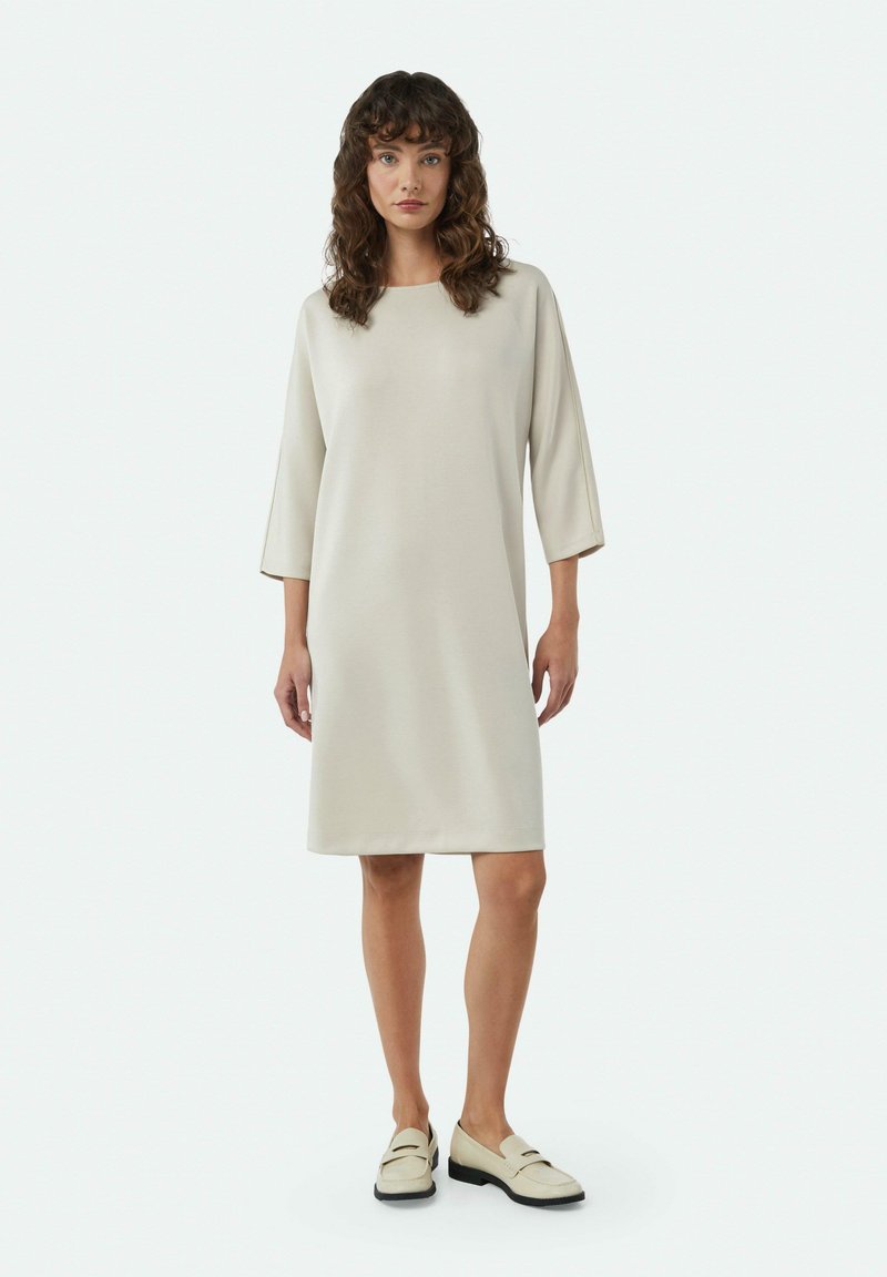 Beige Midi-Kleid mit Dreiviertelärmeln, Rundhalsausschnitt und gerader Silhouette. Tragend mit hellen Slippern. Glatte Textur.