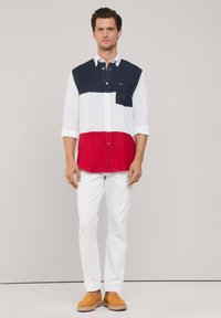 Camisa a bloques de color con secciones en azul marino, blanco y rojo; mangas largas, cuello abotonado, con un bolsillo en el pecho. Combinada con pantalones blancos y zapatos beige.