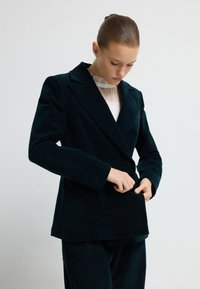 Žena s hladce učesanými vlasy zapíná knoflík na tmavě modrém kordovém blejzru, pod kterým má na sobě bílou průhlednou halenku s vysokým stojáčkem.