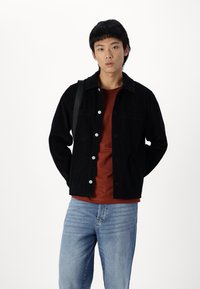 Black corduroy jacket with silver buttons, worn over a rust-colored shirt, paired with light blue jeans, in a neutral background.