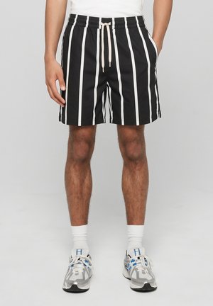 Mand iført sorte shorts med hvide lodrette striber, en hvid snøre, hvide sokker og hvide/grå sneakers, der står mod en ensfarvet baggrund.
