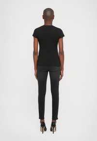 Persoon die met de rug naar de camera staat, gekleed in een aansluitend zwart T-shirt, zwarte skinny jeans en zwarte hoge hakken, tegen een effen achtergrond.