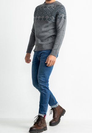 Mann trägt einen grauen Pullover mit Muster, blaue hochgekrempelte Jeans und braune Schnürstiefel, geht vor weißem Hintergrund.
