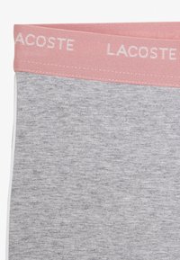 Szary materiał z różowym pasem, na którym powtarza się białe logo tekstowe "LACOSTE", zbliżenie na szczegóły odzieży.