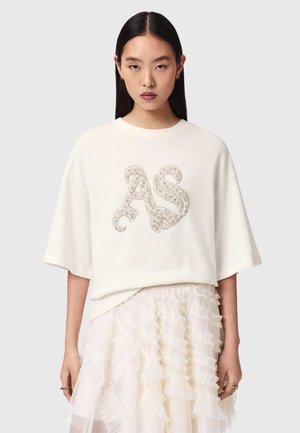 Jonge vrouw met lang donker haar draagt een wit oversized T-shirt met versierde "AS"-letters en een gelaagde geplooide crèmekleurige rok.