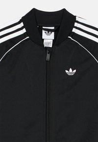 Čierna bunda na zips s rebrovaným golierom, s bielymi pruhmi na ramenách a bielym logom na hrudi. Hladká textúra tkaniny.