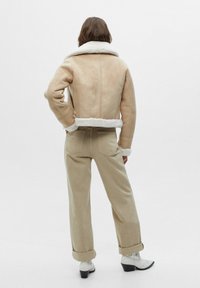 Beige shearlingjas met een brede kraag en omgeslagen zoom, gecombineerd met lichtbeige broek en witte cowboylaarsjes tot aan de enkel. Effen achtergrond.