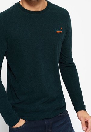 Mężczyzna noszący ciemnozieloną bluzę z długim rękawem i okrągłym dekoltem, z małym haftem na górnej lewej stronie klatki piersiowej, w połączeniu z ciemnoniebieskimi dżinsami.