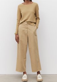 Nainen, jolla on päällään beige ribattupaita, leveälahkeiset beige housut ja kermaiset paksupohjaiset loafertossut, seisoo valkoista seinää vasten.