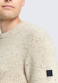 cremefarbener Strickpullover mit strukturiertem Finish, verziert mit mehrfarbigen Sprenkeln und einem schwarzen Etikett-Patch am linken Ärmel.