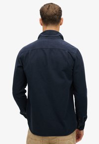Uomo con capelli castani corti che indossa una camicia a maniche lunghe blu scuro e pantaloni beige, rivolto di spalle su uno sfondo bianco semplice.