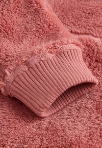 Nærbillede af en pink fleece-ærme med ribstrikket kant på en blød, tekstureret stofoverflade.