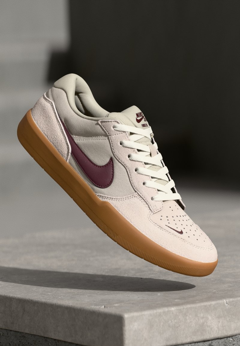 Baskets Nike blanches et beiges avec semelle marron et swoosh grenat, présentées flottant au-dessus d'une surface en béton sur un fond sombre.