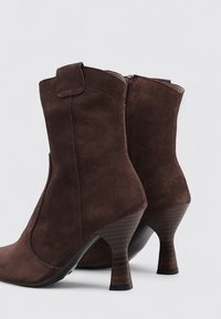 Bottines en suède marron avec un bout rond, un talon haut courbé et une fermeture éclair à l'arrière. Présente une texture lisse et une silhouette classique.