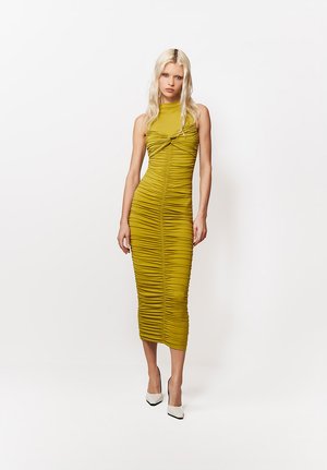 Blondynka w oliwkowej, marszczonej sukience midi bez rękawów i białych, szpiczastych szpilkach, stojąca na tle jednolitego białego tła.