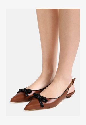 Bruine slingback platte schoenen met spitse neus en zwarte strikaccenten, gedragen op blote voeten tegen een effen achtergrond.