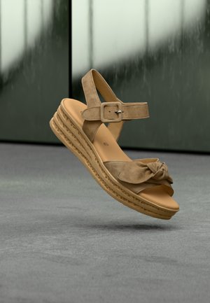 Tan suede wedge sandal with a knotted front strap, adjustable ankle buckle, and layered textured sole. Floating above a gray surface.