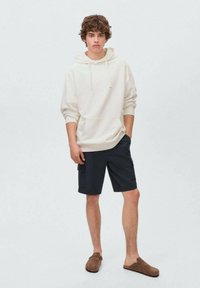 Krämfärgad hoodie med känguruficka, i kombination med mörkblå cargoshorts. Modellen bär bruna slip-on skor. Enfärgad bakgrund.