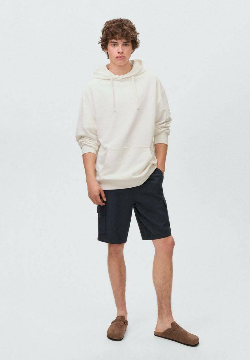 Krämfärgad hoodie med känguruficka, i kombination med mörkblå cargoshorts. Modellen bär bruna slip-on skor. Enfärgad bakgrund.