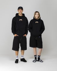 Dos personas llevan sudaderas negras con un logo, pantalones cortos negros y zapatos negros. Las sudaderas tienen bolsillos tipo canguro y cordones.
