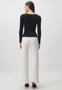 Canottiera nera a maniche lunghe aderente con scollo a giro, abbinata a pantaloni bianchi a gamba larga e scarpe con tacco alto nere, vista da dietro.