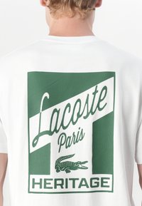 Balta medvilninė marškinėlių su drąsiu žaliu grafiniu spaudiniu ant nugaros, kuriame užrašyta "Lacoste Paris Heritage" ir krokodilo logotipas.