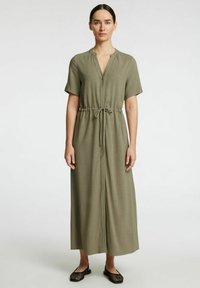 Robe maxi vert olive à manches courtes avec un col en V, taille cintrée et tissu léger. Des chaussures plates noires complètent la tenue.