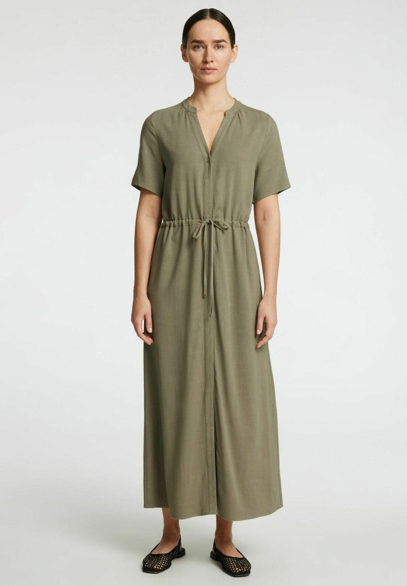Robe maxi vert olive à manches courtes avec un col en V, taille cintrée et tissu léger. Des chaussures plates noires complètent la tenue.