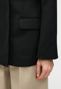 Δεξί χέρι ατόμου που φοράει μαύρο σακάκι και μπεζ παντελόνι, με φακέλο τσέπης σακακιού και μέρος του μανικιού ορατά.