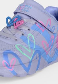 Baskets mauves claires avec des motifs de cœurs colorés, des breloques en forme de cœur sur les lacets et une lanière velcro. Le nom de la marque est visible sur le côté.