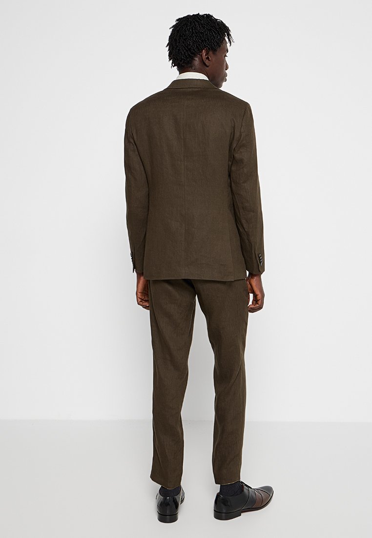 Brauner Leinenanzug mit schmaler Passform, bestehend aus einem einreihigen Sakko und eleganten Hose mit flachem Schnitt. Getragen mit schwarzen Schuhen und minimaler Gestaltung.