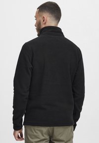 Chaqueta de forro polar negra con un alto cuello, mangas largas y una textura suave. Presenta un dobladillo recto y un detalle de yugo en la parte posterior.