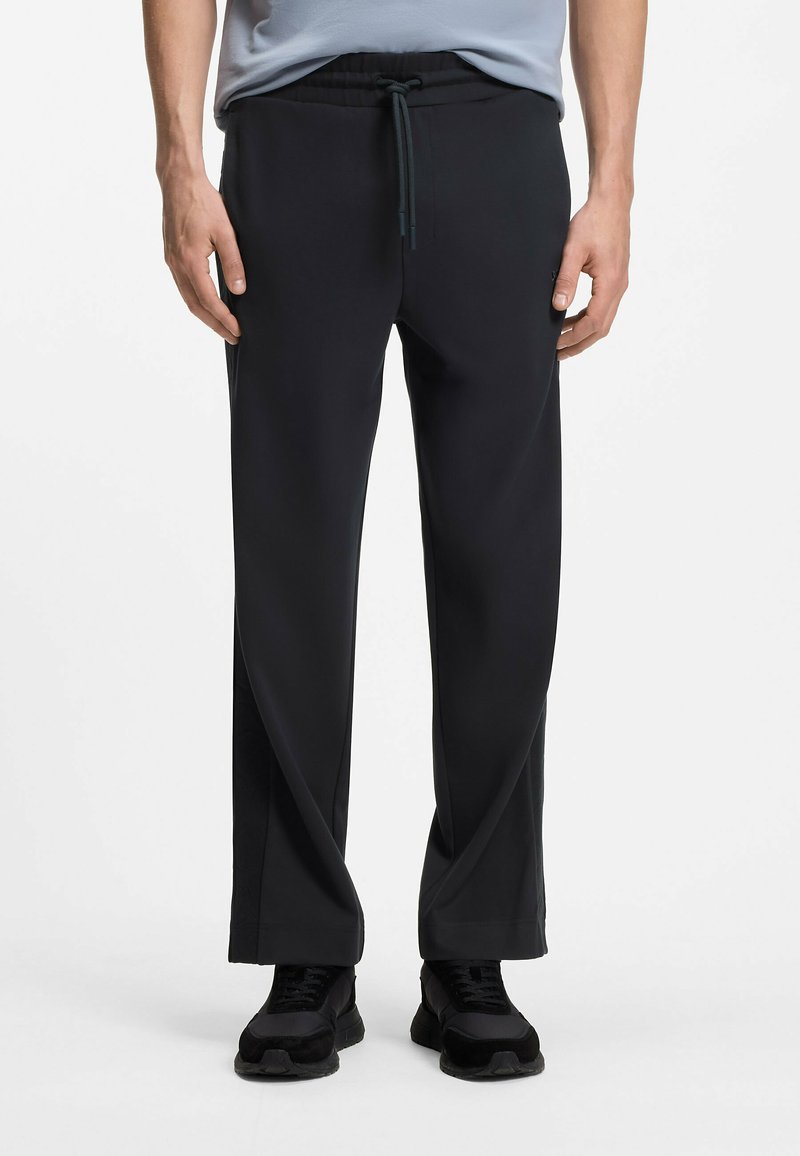 Homme portant un pantalon noir ample avec une taille à cordon et des baskets noires, debout sur un fond blanc.