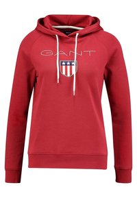 Sudadera con capucha de algodón roja con bolsillo tipo canguro. Cuenta con capucha ajustable con cordones, logo bordado "GANT" y un escudo con franjas rojas y azules.