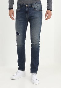 Slim-fit blekta blå jeans med en slitna lappar på vänstra låret, som kombineras med vita sneakers och en randig långärmad skjorta.