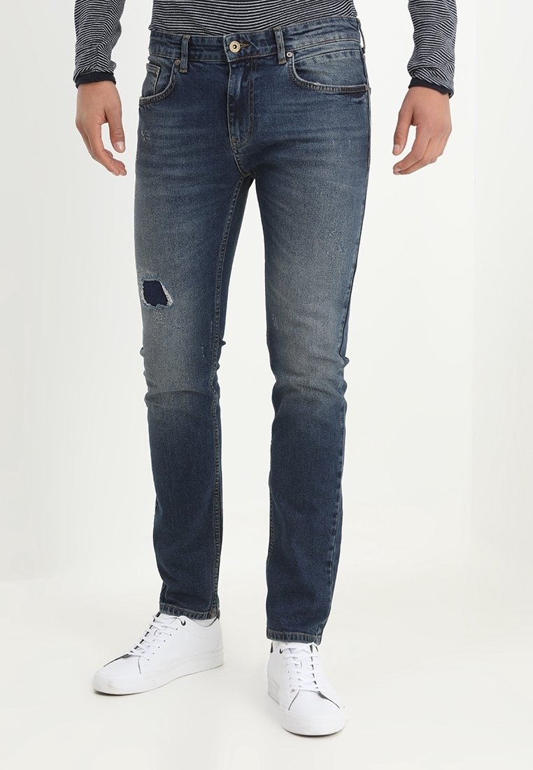 Slim-fit blekta blå jeans med en slitna lappar på vänstra låret, som kombineras med vita sneakers och en randig långärmad skjorta.