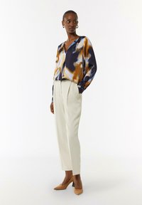 Seidenbluse mit einem mehrfarbigen Muster in Navy, Orange und Beige; kombiniert mit cremefarbenen, maßgeschneiderten Hosen und braunen, hochhackigen Schuhen.