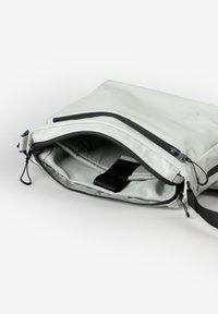Lichtgrijze tas met een gladde textuur, voorzien van een zwarte rits en band, geopend om een zachte binnenvoering en een zwarte band aan de binnenkant te tonen.