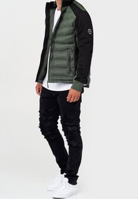 Zielona pikowana kurtka puchowa z czarnymi rękawami, zamkiem błyskawicznym i ściągaczami na rękawach. Do tego podarte czarne spodnie i białe trampki.