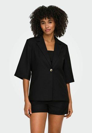 Vrouw met krullend haar die een zwarte korte mouwen blazer met één knoop en bijpassende zwarte shorts draagt tegen een effen achtergrond.